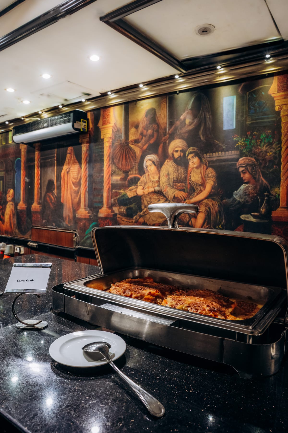 A buffet chafing dish with carrot gratin sits before an ornate mural in the ship's restaurant.